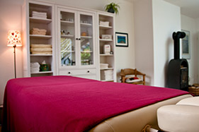 Behandlungsliege mit roter Decke, dahinter ein heller Schrank und ein Kaminofen